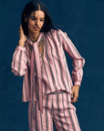 A woman with long dark hair smiles and touches her hair while wearing the Xirena Sabina Top—a drapey pink and black vertical striped blouse—paired with a matching scarf and pants, set against a dark blue background.