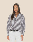 A woman wears the Xirena Monroe Shirt, a loose black-and-white striped cotton top, tucked into white pants with a black belt, standing against a plain beige background.