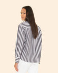 A woman with long, straight brown hair models the Xirena Monroe Shirt—a button-front, black and white striped cotton top—paired with white pants, standing against a beige background and looking over her shoulder.