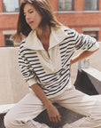 A woman with long brown hair sits outdoors in sunlight, wearing the Kat Sweatshirt by Xirena—a cream and navy striped cotton zip-collar top—paired with light pants. A brick building can be seen in the background.