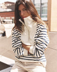 A woman with long brown hair sits outdoors wearing the Xirena Kat Sweatshirt, a striped cotton top with a high collar, paired with light pants. She looks at the camera neutrally, with urban buildings in the background.