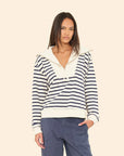 A woman stands against a beige background wearing the Xirena Kat Sweatshirt—a navy and white striped cotton top with a wide collar and cream trim—paired with blue pants, one hand in her pocket.