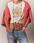 Person wearing a white knotted t-shirt with a golden sun "Follow the Sun" graphic, red cardigan draped over shoulders, beaded necklaces and bracelets, the Stevie Belt by Xirena, and blue jeans. Hands are tucked in pockets.