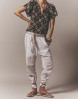 A person poses in a black patterned blouse, loose white pants with the Stevie Belt by Xirena, ankle-tied espadrille sandals, and several bracelets against a neutral background. Their head is not shown in the image.
