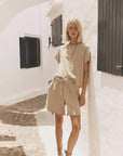 A blonde woman stands against a white wall in the sun, wearing the Xirena Rocco Short and matching top. She completes her look with sandals, a long necklace, and dark window shutters in the background.