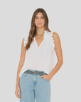 A woman wearing the Xirena Bexie Top, a white cotton gauze blouse with ruffle details, stands against a plain light gray background. She pairs it with light blue jeans, a beaded belt, and a green necklace.
