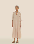In front of a beige backdrop, an individual models a Xirena dress fashioned from soft cotton poplin. The Boden dress features rolled-up sleeves, and they accentuate their look with braided hair, layered necklaces, and intricately laced-up sandals.