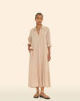 A person stands against a light beige background, wearing the Boden Dress by Xirena, a long button-front tailored shirtdress in light tan with sleeves rolled to the elbows. They have braided hair, layered necklaces, and strappy sandals.