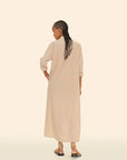 A person with braided hair stands facing away, wearing the Boden Dress, a long, tailored piece by Xirena, paired with strappy sandals. The background is a simple off-white color.