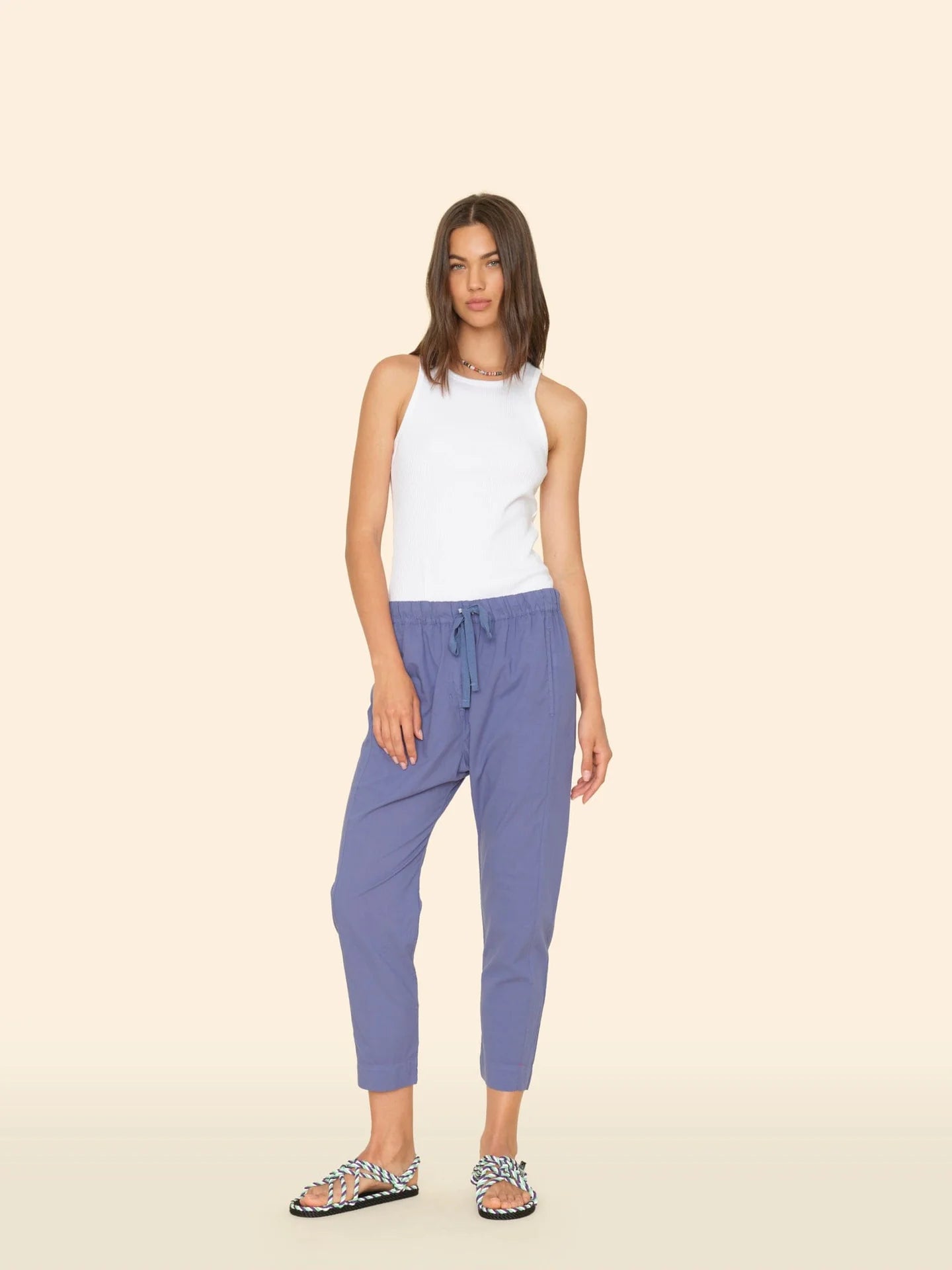 A woman with straight, shoulder-length hair stands against a beige background wearing the Xirena Draper Pant—blue joggers with a drawstring waist and side seam pockets—paired with a white sleeveless top and patterned slide sandals.