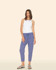 A woman with straight, shoulder-length hair stands against a beige background wearing the Xirena Draper Pant—blue joggers with a drawstring waist and side seam pockets—paired with a white sleeveless top and patterned slide sandals.