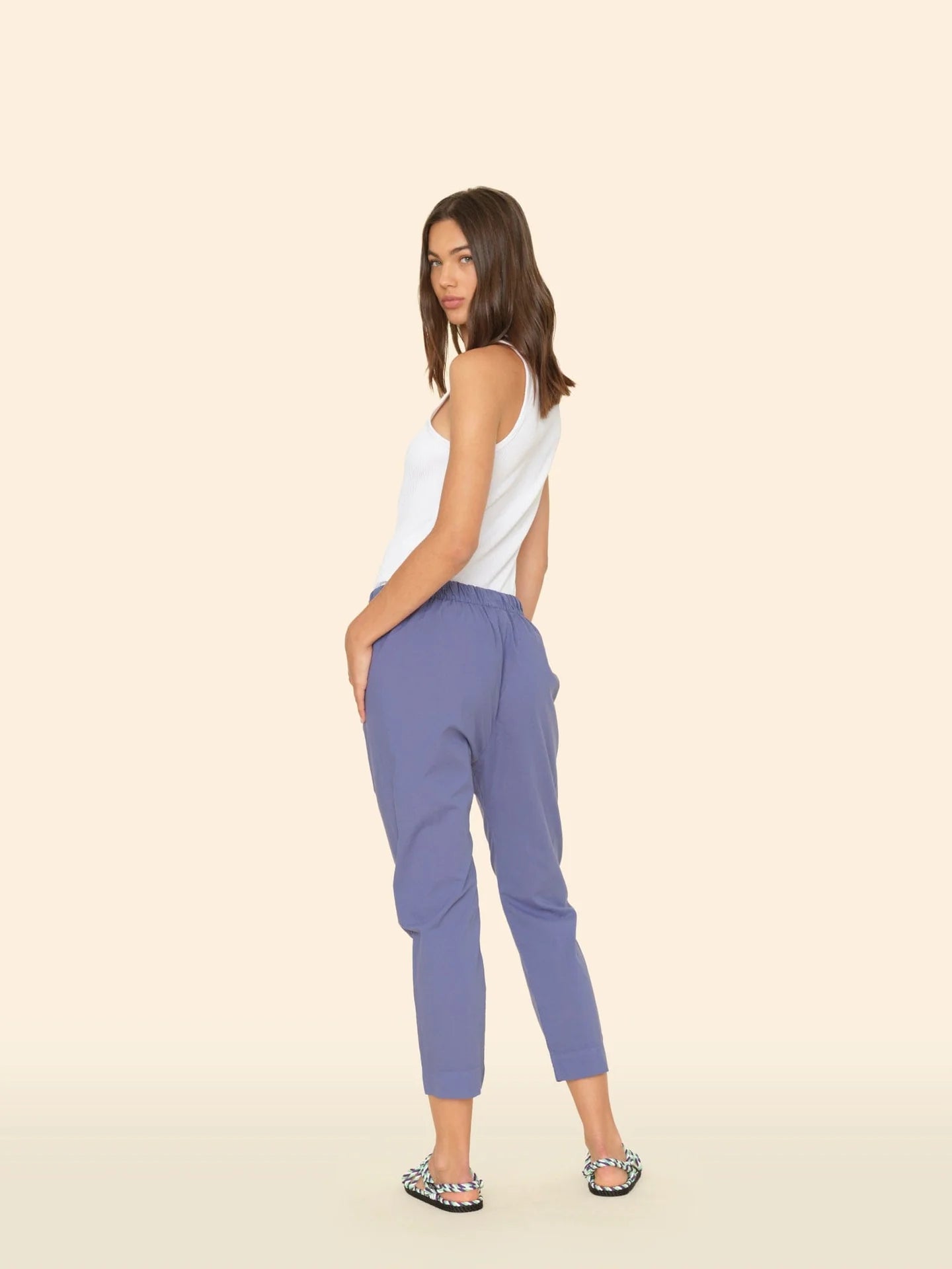 A woman with shoulder-length brown hair wears the Xirena Draper Pant in blue lightweight cotton chambray, featuring side seam pockets, styled with a white sleeveless top and patterned sandals, standing sideways against a beige background.