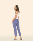 A woman with shoulder-length brown hair wears the Xirena Draper Pant in blue lightweight cotton chambray, featuring side seam pockets, styled with a white sleeveless top and patterned sandals, standing sideways against a beige background.