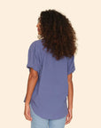 A woman with curly hair stands facing away, wearing the Xirena Channing Shirt S25 in purple and light blue jeans against a plain beige background.