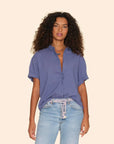 A woman with curly hair wears the Xirena Channing Shirt S25 in lightweight blue chambray, styled with light blue jeans and a woven belt, channeling a "boy meets girl" vibe against a plain beige background.