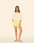 A woman stands against a light background wearing a yellow and white striped long-sleeve shirt, Shayne Sweatshorts by Xirena, beige sandals, and maintains a neutral expression with relaxed hands at her sides.