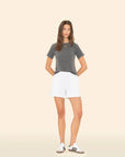 A woman stands against a plain background, wearing an Xirena gray t-shirt and Shayne Sweatshorts featuring side seam pockets. She completes the look with white sneakers accented by black stripes, her hands behind her back, and her hair cascading casually.