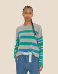 A person wearing the ultra-soft Xirena Lela Sweater, featuring vibrant blue and green stripes, stands against a beige background. They have long braided hair and are dressed in blue jeans paired with a light blue belt.