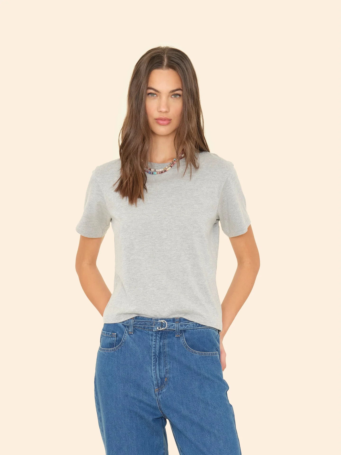 A woman with long hair wears a Georgy Tee by Xirena, styled with blue jeans and a colorful beaded necklace against a neutral backdrop. The gray, boxy-fit tee complements her relaxed look, as her hands rest casually in her pockets.