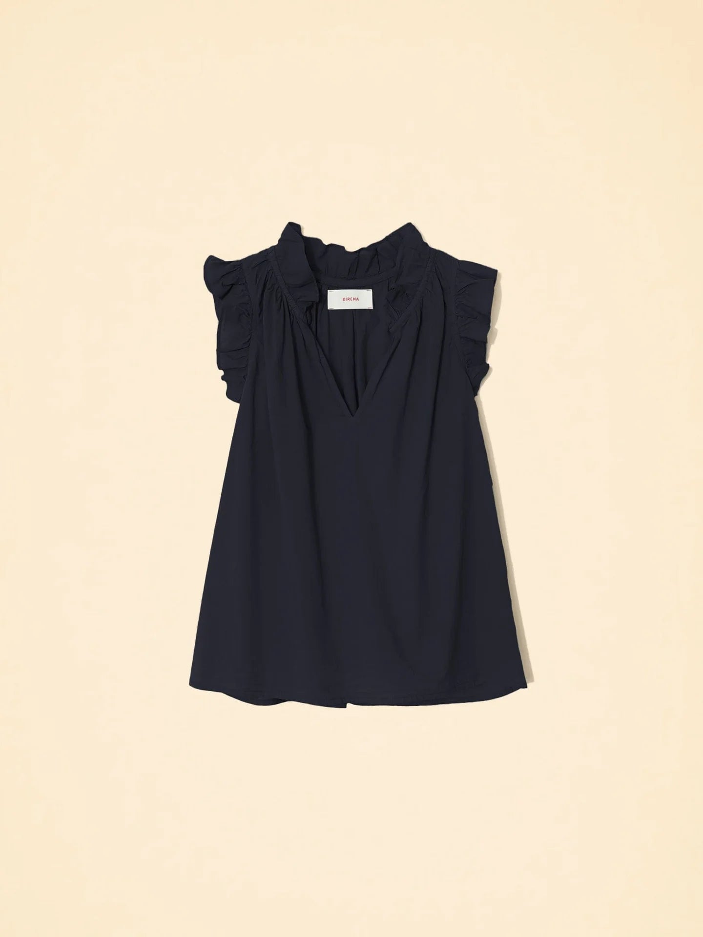 The Bex Top by Xirena is a sleeveless black blouse with a deep V-neck and elegant ruffle shoulders. Made from lightweight cotton poplin, it flows naturally and stands out beautifully against a light beige background.