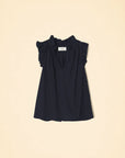 The Bex Top by Xirena is a sleeveless black blouse with a deep V-neck and elegant ruffle shoulders. Made from lightweight cotton poplin, it flows naturally and stands out beautifully against a light beige background.