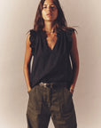 A woman with long hair dons a sleeveless Bex Top by Xirena and olive cotton poplin pants, hands in pockets. She accessorizes with a necklace and bracelets, confidently facing the camera against a neutral backdrop.