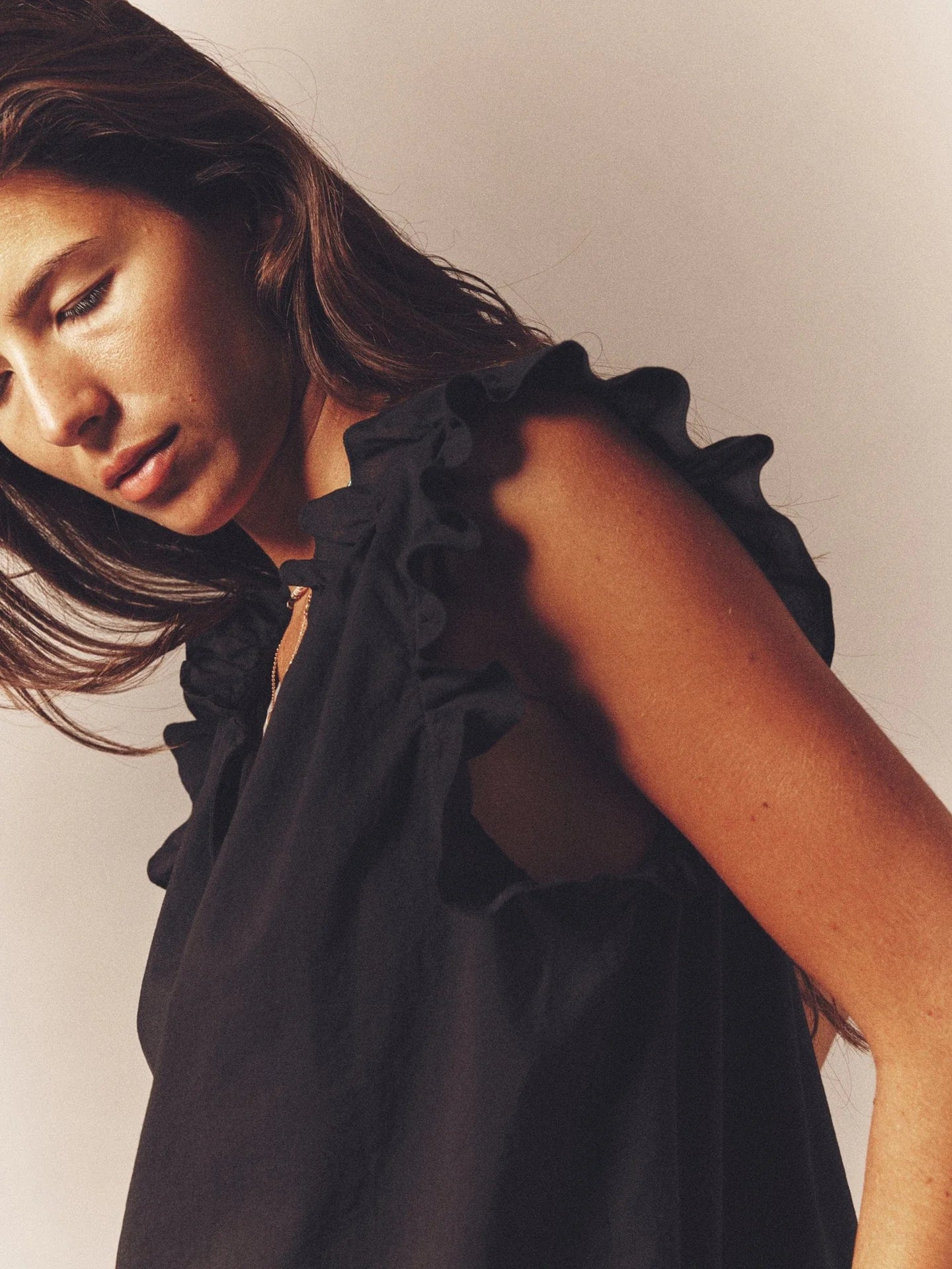 A woman with long brown hair gazes downward pensively, wearing Xirena's Bex Top, a sleeveless black piece featuring ruffle detailing along the V-neck. Set against a plain background, warm lighting accentuates the fabric's texture and her contemplative expression.