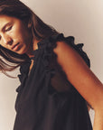 A woman with long brown hair gazes downward pensively, wearing Xirena's Bex Top, a sleeveless black piece featuring ruffle detailing along the V-neck. Set against a plain background, warm lighting accentuates the fabric's texture and her contemplative expression.
