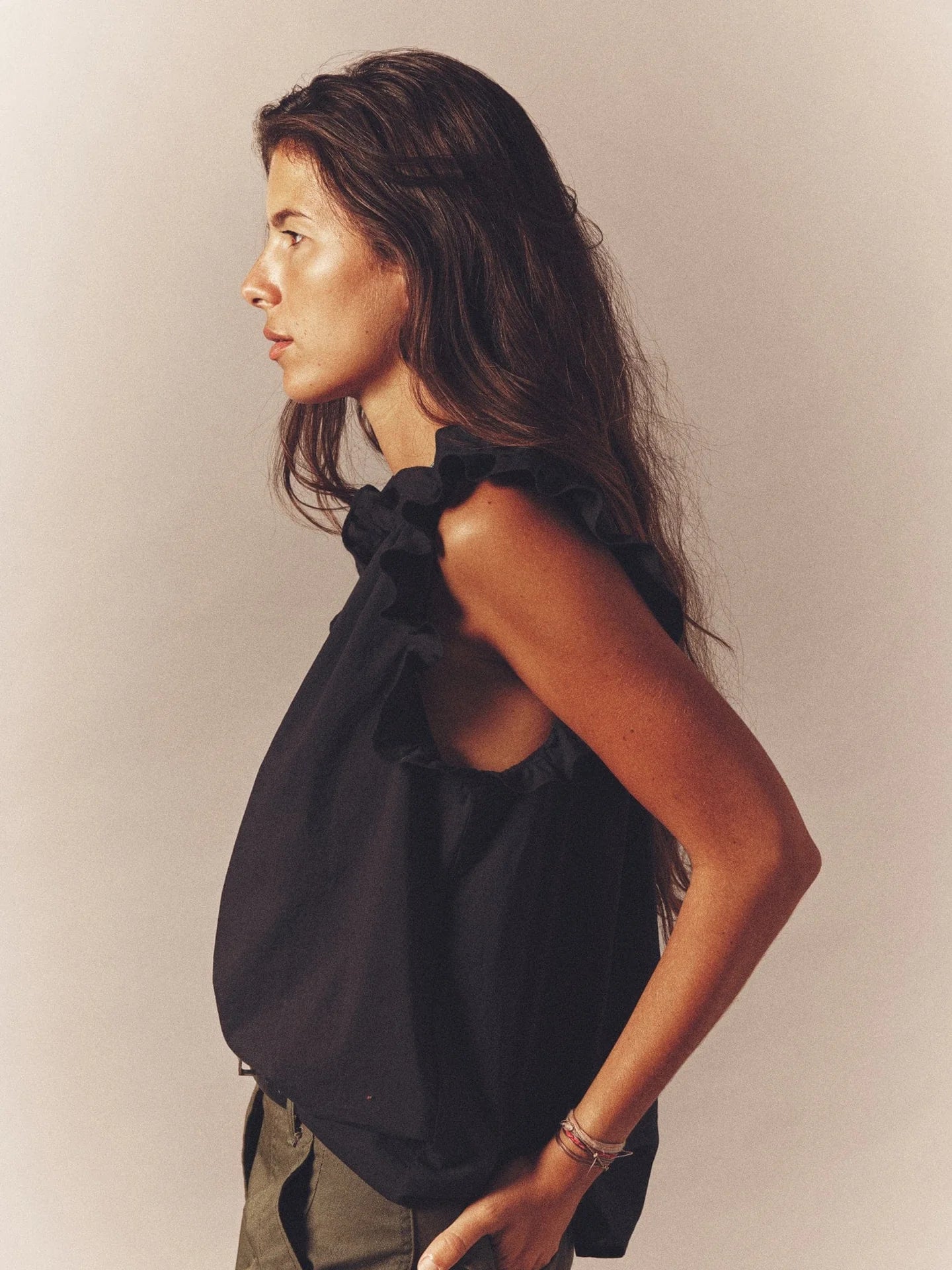 A woman with long dark hair wears the sleeveless black Bex Top by Xirena, crafted from soft cotton poplin with ruffle detailing and a subtle V-neck. She gazes left against a plain background, her expression neutral.