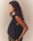 A woman with long dark hair wears the sleeveless black Bex Top by Xirena, crafted from soft cotton poplin with ruffle detailing and a subtle V-neck. She gazes left against a plain background, her expression neutral.