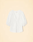The Jules Top by Xirena is a white cotton poplin blouse featuring a loose fit, three-quarter bubble sleeves, a gathered neckline, and a V-shaped front slit, shown against a light beige background.