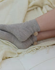Someone wears Unified’s Winter Pom Pom Socks in cozy gray, resting their legs on a cream-colored blanket draped over a white surface.