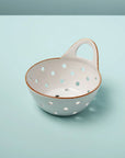 The Yara Colander Basket by Be Home is a handcrafted stoneware bowl featuring large circular holes, a single handle, and a rustic unglazed rim, shown resting on a light blue surface for added charm.