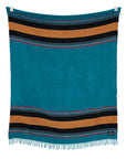 The Sackcloth & Ashes/Yellowstone Teal blanket by Sackcloth & Ashes, made from recycled materials, displays turquoise with black and gold stripes at the top and bottom, small fringe edges, and is shown hanging against a white background.