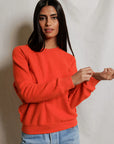 A person with long dark hair wears the Perfectwhitetee Ziggy Reverse LS Sweatshirt in bright orange cotton fleece (shrunken fit) and light blue jeans, standing against a plain light background, looking at the camera and adjusting one sleeve.