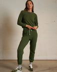 A woman stands indoors against a plain wall, wearing the Perfectwhitetee Ziggy Reverse LS Sweatshirt in olive green with matching jogger pants, gray socks, and green and white Nike sneakers. She looks confidently at the camera, hands on her waist.