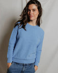A woman with long brown hair wears the Perfectwhitetee Ziggy Reverse LS Sweatshirt in light blue and blue jeans, standing with hands in pockets and smiling softly in front of a plain, light-colored draped backdrop.