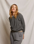 A person with long hair, wearing a cozy Ziggy Reverse Fleece LS Crew Sweatshirt by Perfectwhitetee and matching pants, stands against a plain light-colored backdrop, smiling softly.