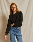 A person with long red hair, wearing the Perfectwhitetee Ziggy Reverse LS Sweatshirt in black and blue jeans, stands before a beige fabric backdrop, looking relaxed with one arm bent.