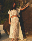 A woman stands barefoot on a sunny beach, leaning against a rock and wearing the DOEN Zinnia Skirt paired with a matching white short-sleeved crop top. Warm sunlight adds to the relaxed, elegant look.