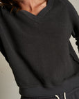 Someone wearing the Perfectwhitetee Zuri Reverse V Neck sweatshirt and matching drawstring pants in textured dark gray; the cropped image shows only their midsection, partial face, and relaxed raglan sleeves.