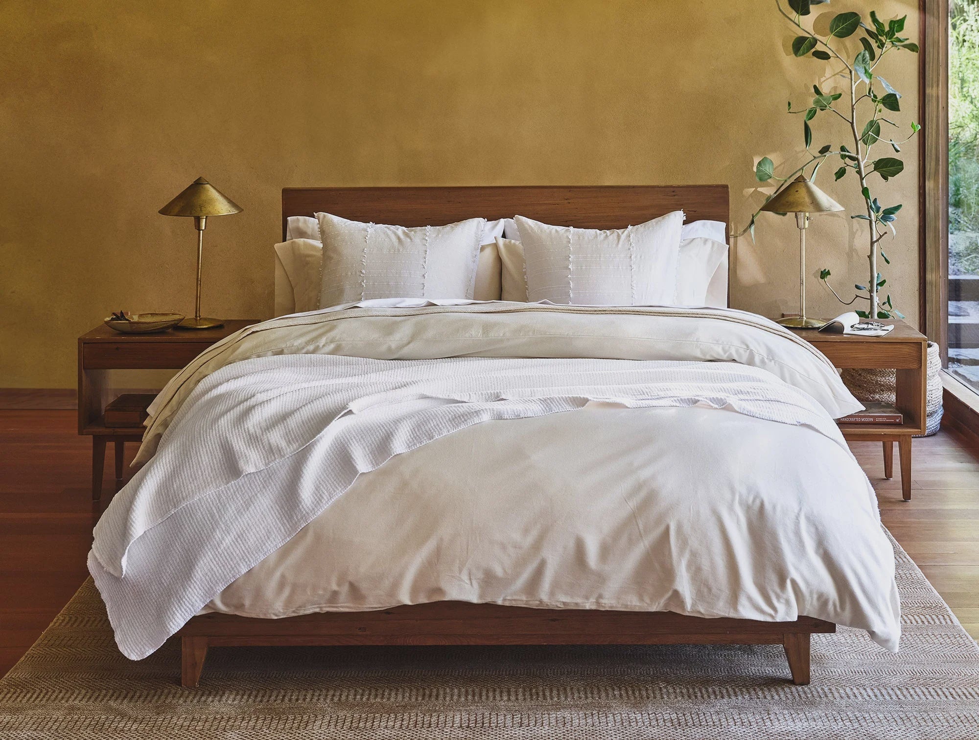 A neatly made bed with white Coyuchi Inc bedding and wooden frame in a serene bedroom with gold walls, side tables with lamps, and a potted plant in a Scottsdale Arizona bungalow.
