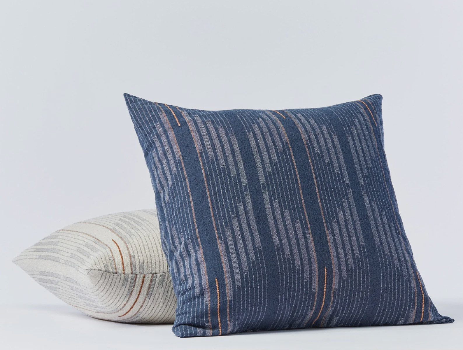 Two Morel Euro Sham Moon. Blue against a plain background; one upright with deep blue and subtle stripe design, and one leaning with a Coyuchi Inc Bungalow-style cream and multicolored curved stripe pattern.