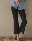 A person dressed in a striped blue shirt and Donni black Rib Kick Flare trousers is standing on a woven rug in a Scottsdale, Arizona bungalow, with hands clasped in front. Only the lower torso and legs are visible.