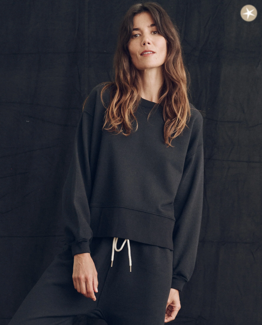 A woman in the casual dark grey "The League" sweatshirt by The Great Inc. and pants stands confidently against a black textured backdrop, her brown hair loose and wavy, reminiscent of the relaxed vibe of a Scottsdale, Arizona bungalow.