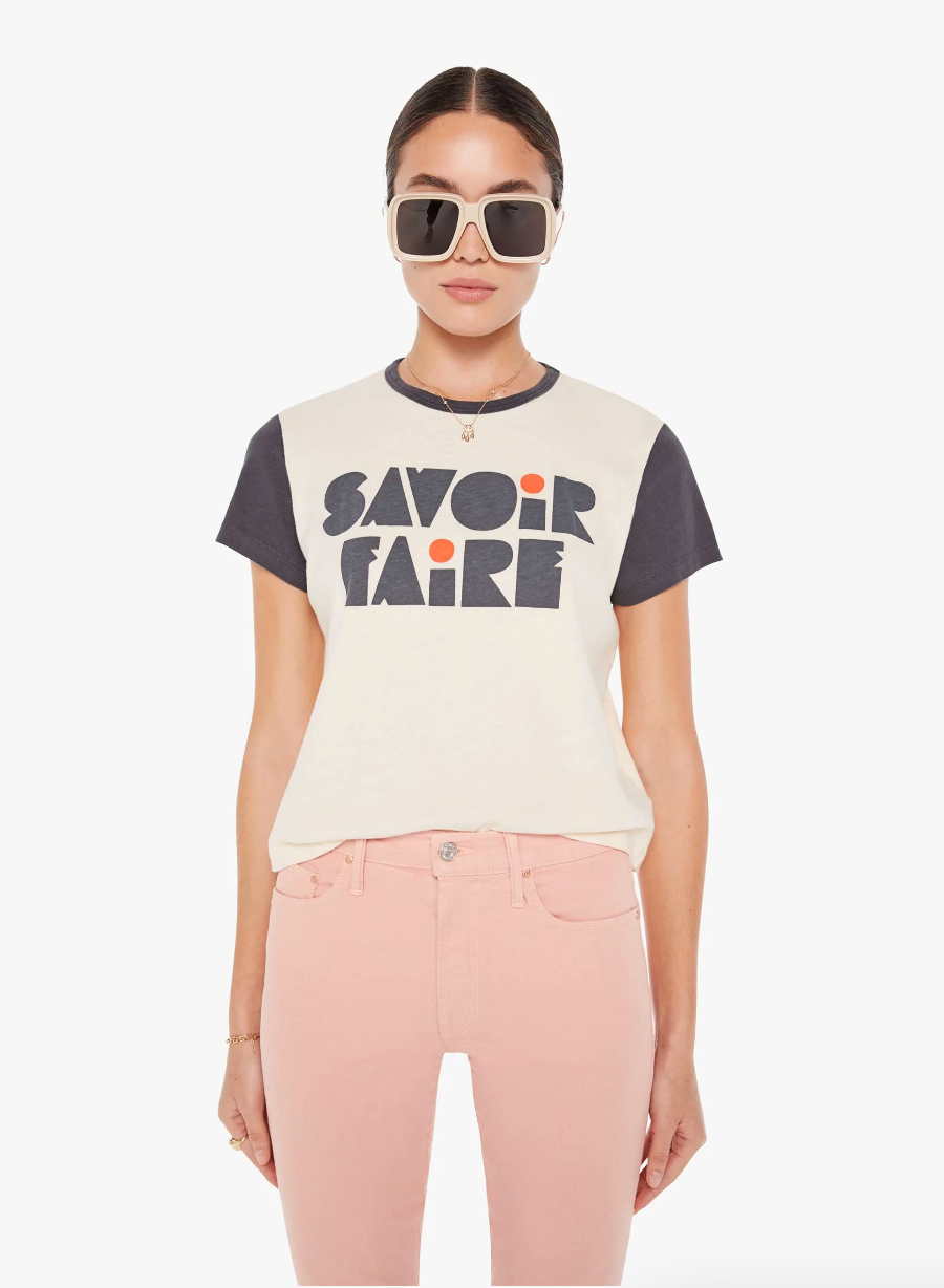 A woman in sunglasses wearing a Mother Goodie Goodie ringer Savoir Faire T-shirt and light pink jeans, standing against a white bungalow background.