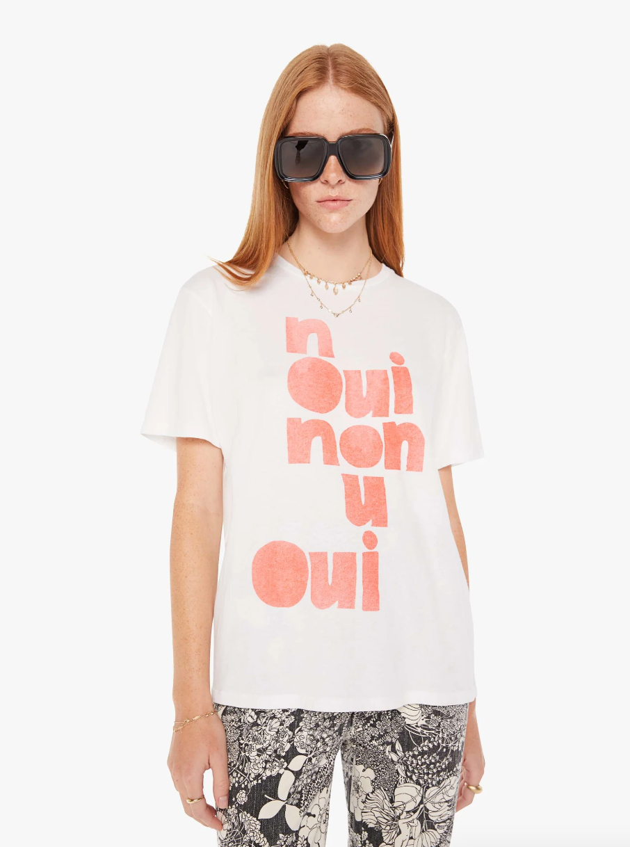 A woman in a white T-shirt with "the Rowdy Oui Non" printed in large pink letters, paired with floral pants reminiscent of Scottsdale Arizona, wearing oversized sunglasses and a pearl necklace by Mother.