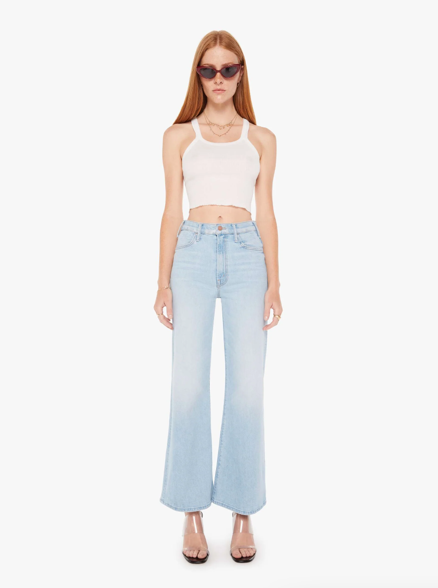 A woman wearing a white crop top, light blue wide-leg jeans, and clear high heels accessorizes with sunglasses and stands against a plain white background in Scottsdale, Arizona is wearing the Mother Hustler Roller Flood Cherie Cherie.