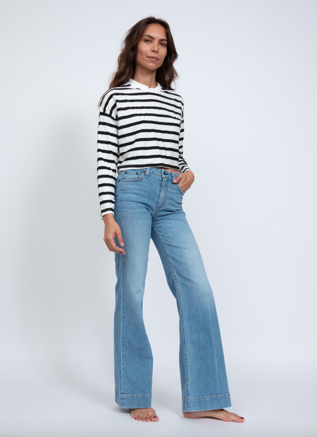 A person stands against a white background wearing an ASKK printed cropped long sleeve and wide-leg blue jeans. Barefoot with loose long hair, their effortless style embodies Los Angeles' laid-back vibe.