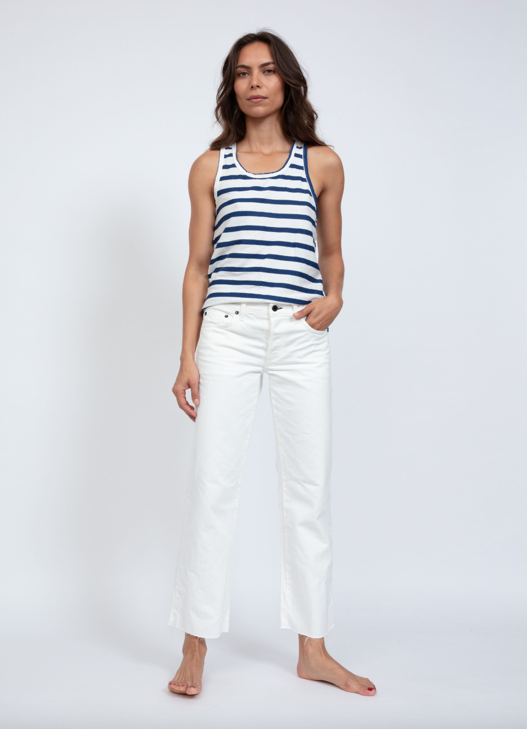A woman stands confidently in a studio in Scottsdale, Arizona, wearing an ASKK striped tank top and white jeans, hands slightly tucked into pockets, on a plain background.